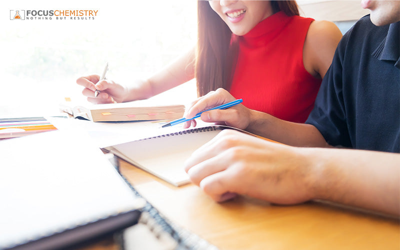 A bright, close-up shot of two individuals sitting side-by-side, focusing on a shared open textbook and a spiral notebook.