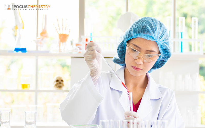 Scientist in lab coat and goggles pipetting red liquid into test tube amid colorful glassware and equipment.